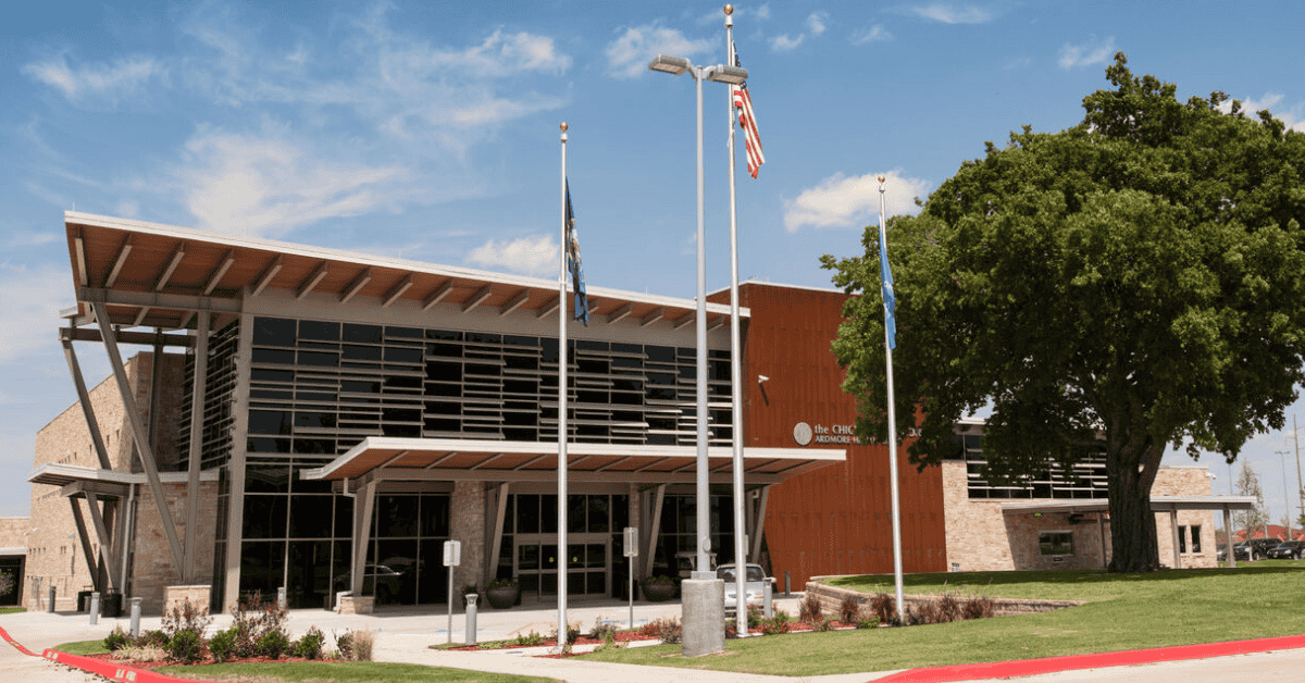 Chickasaw Nation Department of Health facility in Ardmore, Oklahoma, with angular metal roof design and stone exterior. Main entrance includes American and tribal flags amid mature landscaping.