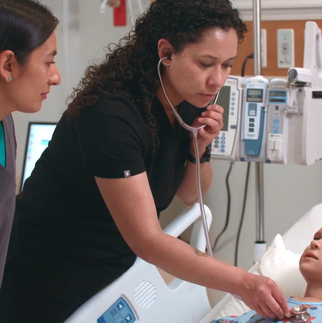 A healthcare professional with curly hair in black scrubs uses a stethoscope to examine a patient in a hospital bed. Medical equipment and monitors are visible in the background, while another healthcare worker observes nearby.