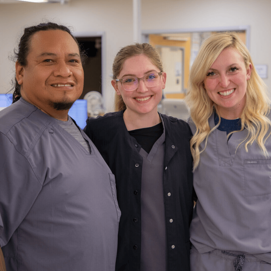 Three nurses - one male and two female - stand closely together in a hospital hallway. They are wearing scrubs and smiling warmly at the camera, conveying a sense of teamwork and camaraderie.