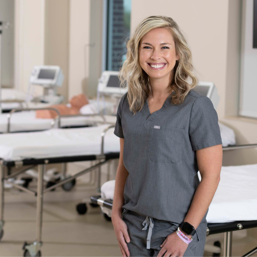 A smiling nurse wearing gray scrubs stands in the foreground. In the blurred background, hospital gurneys are visible, with nursing simulation equipment resting on one of them.